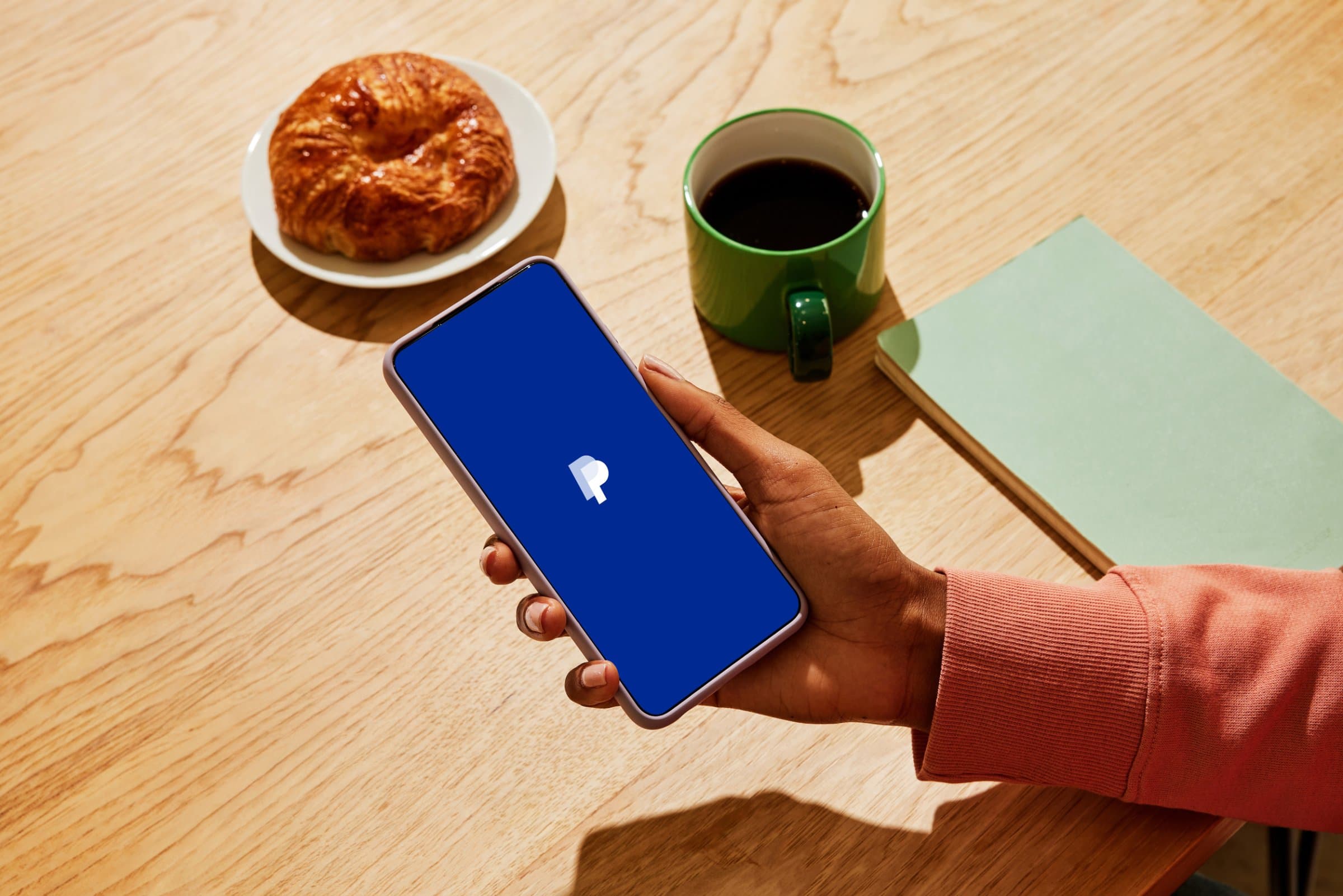 Hand holding a phone showing the PayPal app on a cafe table